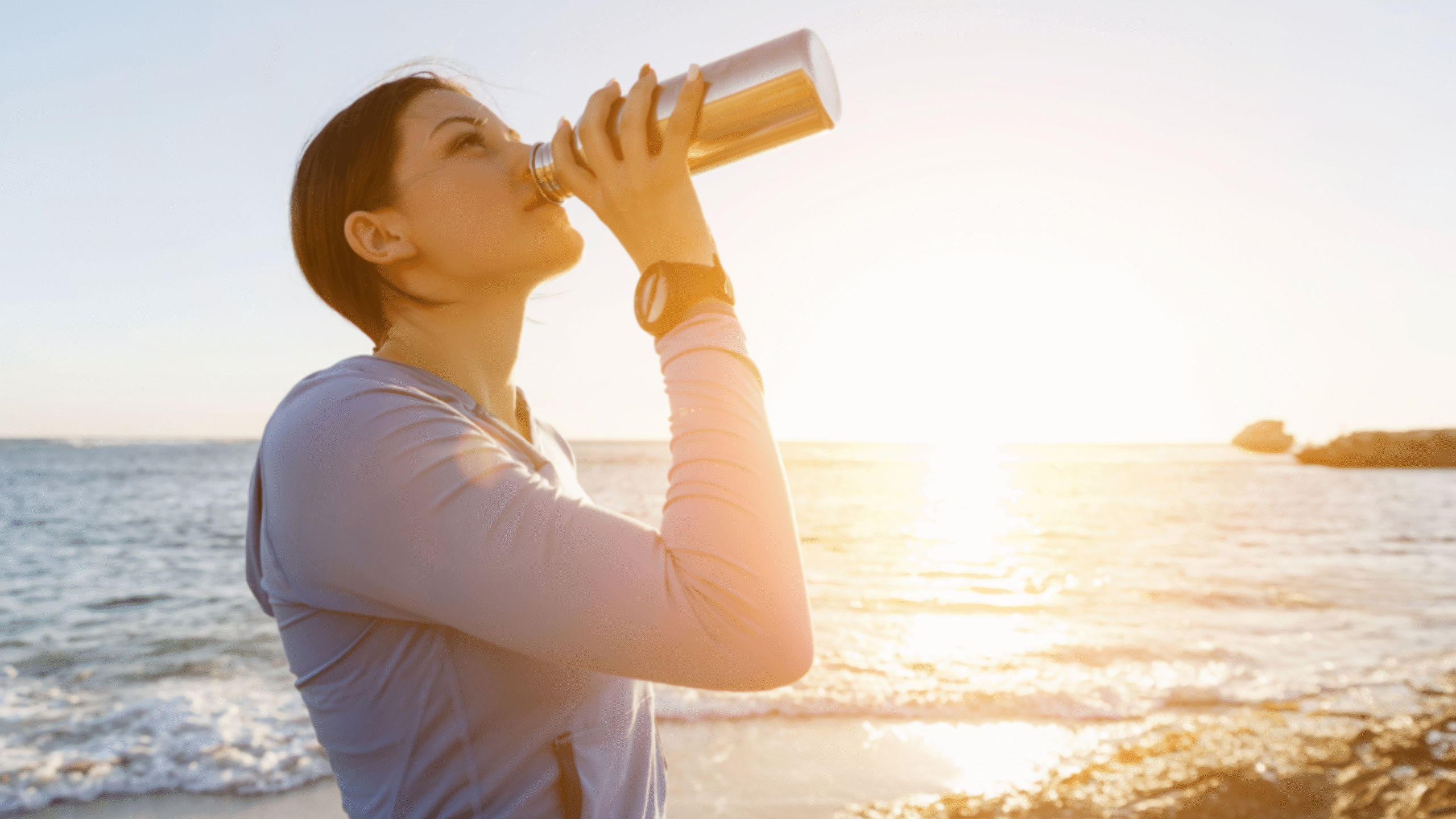 woman drinks water on the beach to stay hydrated